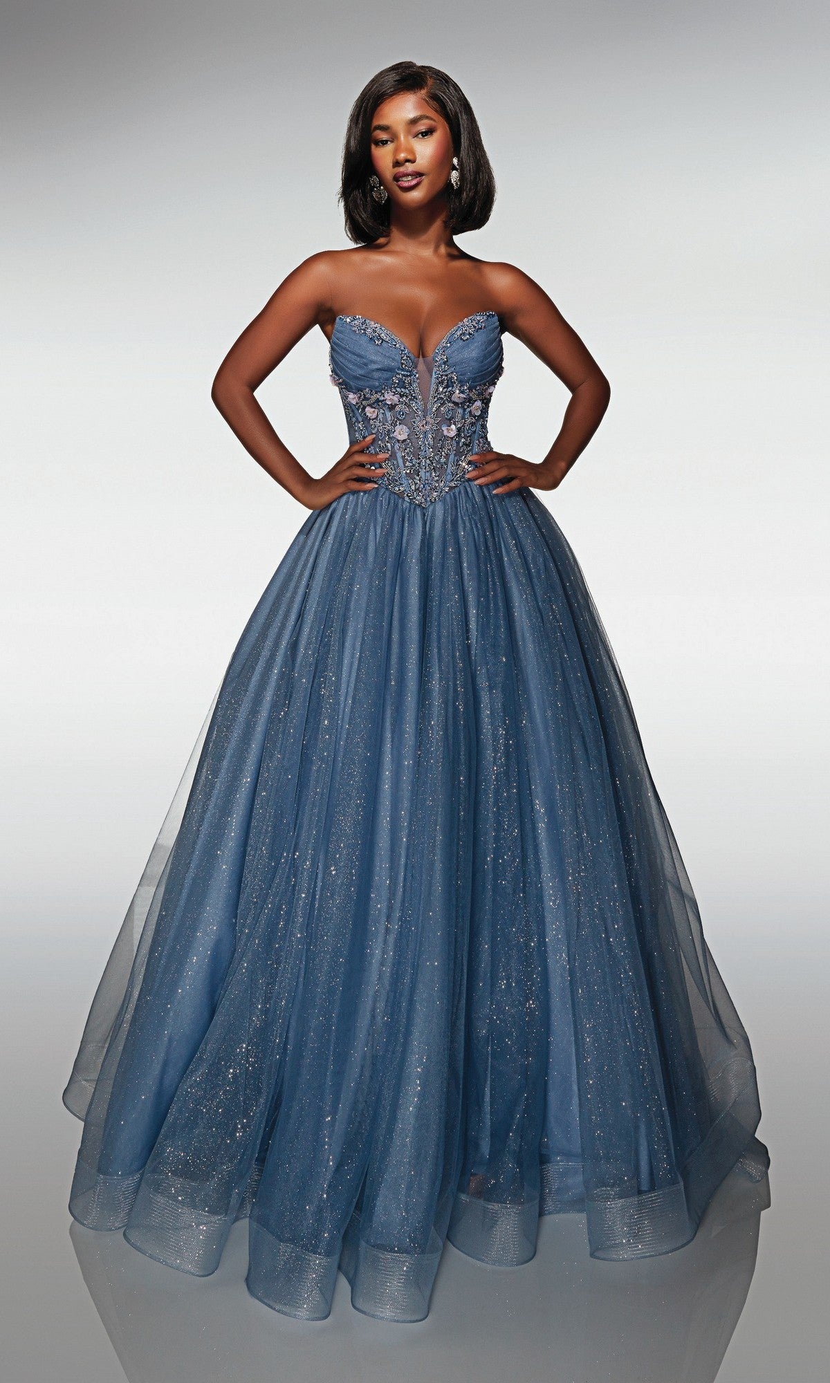 A woman stands with hands on hips, wearing the Alyce 62009 long prom dress featuring a strapless corset bodice, blue 3D flower appliquรฉ, and a full sparkling glitter tulle ball gown skirt.